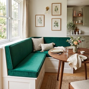 May include: An L-shaped corner bench with deep green velvet upholstery and a white wooden base, paired with a round wooden dining table, two decorative throw pillows, and a floral centerpiece in a ceramic vase set in a kitchen area.