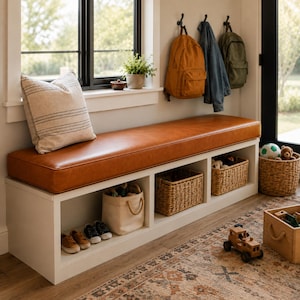 May include: A white storage bench with a long, smooth, tan leather-like cushion on top. The base features three open cubbies containing woven baskets and a canvas bin. A beige throw pillow with thin horizontal stripes sits on the left side.