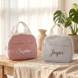 May include: Two personalized lunch bags with white handles. One is red with white stripes and the name "Sophia" embroidered on the front. The other is light blue with white stripes and the name "Jasper". Both bags are on a wooden table.