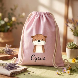 Puede incluir: Una bolsa rosa con cordón con una ilustración de nutria de dibujos animados. La nutria es marrón y blanca con una cara sonriente. El nombre "Cyrus" está impreso en escritura negra debajo. La bolsa tiene cordones color crema.