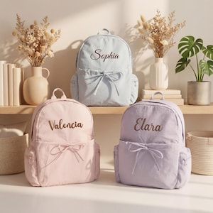Peut inclure: Trois sacs à dos en bleu clair, rose pâle et lavande, chacun doté d'une poche avant avec un nœud coulissant et des noms brodés Sophia, Valencia et Elara. Ces sacs possèdent des poches latérales et des poignées supérieures au design épuré.