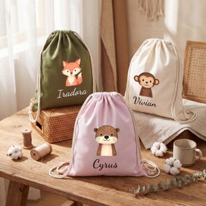 Puede incluir: Tres bolsas con cordón en verde oliva, rosa claro y crema. Cada bolsa presenta una ilustración de animal de dibujos animados y un nombre: un zorro con "Iradora", una nutria con "Cyrus" y un mono con "Vivian". Las bolsas tienen cordones blancos.