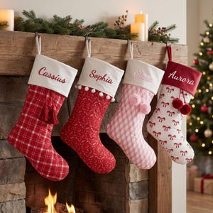 Puede incluir: Cuatro calcetines de Navidad colgados en fila. El primero es de cuadros rojos con puño blanco bordado con Cassius y una borla roja. El segundo es rojo con puño blanco bordado con Sophia y pompones. El tercero es de cuadros rosas con puño blanco bordado con Elara y pompones. El cuarto es blanco con lazos rojos, puño rojo bordado con Aurora y pompones rojos.