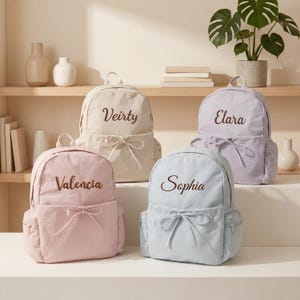 Pode incluir: Quatro mochilas em cores pastel: bege, lavanda, rosa claro e azul claro. Cada mochila possui um bolso frontal com um laço decorativo e é personalizada com nomes bordados: Veirty, Elara, Valencia e Sophia.