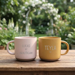 Può includere: Due tazze in ceramica con finitura maculata. La tazza a sinistra è rosa chiaro con il testo LUKAS, un simbolo di cuore e BRAUTIGAM. La tazza a destra è giallo senape con il testo TEYLA e un cuore. Entrambe le tazze hanno un manico arrotondato.