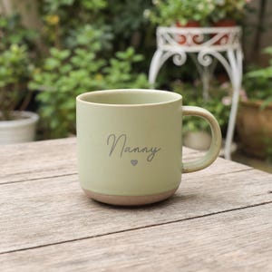 Può includere: Una tazza in ceramica verde salvia chiaro con finitura maculata e un manico liscio. La parola Nanny è stampata in corsivo grigio scuro sul davanti, sopra una piccola icona a forma di cuore grigio. La base della tazza è in argilla non smaltata.