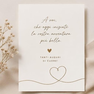Può includere: Una carta color crema con una scrittura elegante in inchiostro marrone. Il testo recita: "A voi, che oggi iniziate la vostra avventura più bella." Sono presenti anche un piccolo cuore e le parole "TANTI AUGURI DI CUORE!". Un delicato contorno a forma di cuore è disegnato in basso.