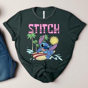 May include: Dark green t-shirt featuring the word "STITCH" in pink block letters above a cartoon character surfing. The character is blue with large ears and is riding a yellow and red surfboard. Palm trees and a sun are also in the design.