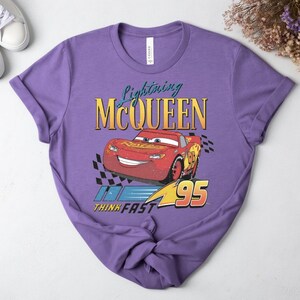 May include: A lilac t-shirt with a graphic of Lightning McQueen from the Cars film. The design includes the text "Lightning McQueen," a red race car, and the words "Think Fast 95."