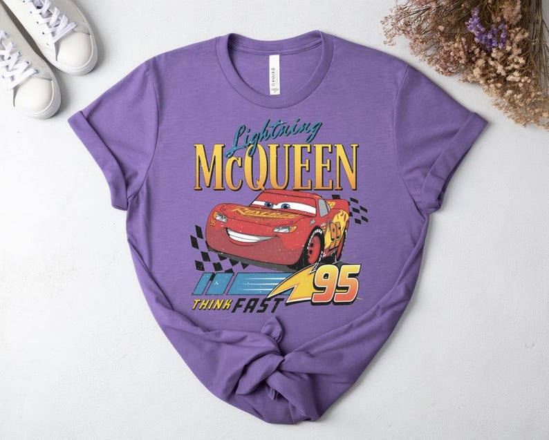 May include: A lilac t-shirt with a graphic of Lightning McQueen from the Cars film. The design includes the text "Lightning McQueen," a red race car, and the words "Think Fast 95."