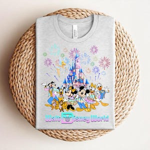 Może przedstawiać: Jasnoszara koszulka z kolorową grafiką postaci Disneya, zamkiem i fajerwerkami. Tekst "Walt Disney World" jest wyświetlany czcionką gradientową na dole. Koszulka jest złożona na tkanej powierzchni.