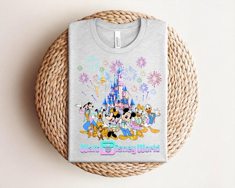 Może przedstawiać: Jasnoszara koszulka z kolorową grafiką postaci Disneya, zamkiem i fajerwerkami. Tekst "Walt Disney World" jest wyświetlany czcionką gradientową na dole. Koszulka jest złożona na tkanej powierzchni.