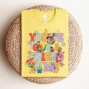 May include: Yellow t-shirt with the text "You've Got a Friend in Me" in a retro font, accompanied by cartoon characters from the Toy Story film. The design includes Woody, Buzz Lightyear, and other characters. The shirt is folded on a woven, circular surface.
