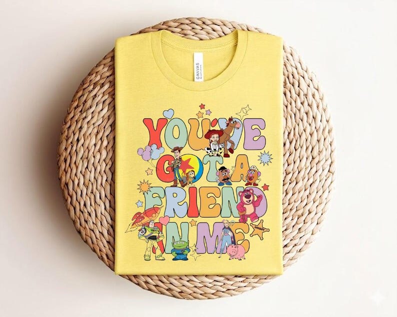 May include: Yellow t-shirt with the text "You've Got a Friend in Me" in a retro font, accompanied by cartoon characters from the Toy Story film. The design includes Woody, Buzz Lightyear, and other characters. The shirt is folded on a woven, circular surface.
