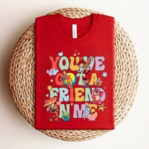 May include: Red t-shirt with the phrase "You've Got a Friend in Me" in colourful, retro lettering. The shirt features cartoon characters. The t-shirt is displayed on a woven, circular mat.