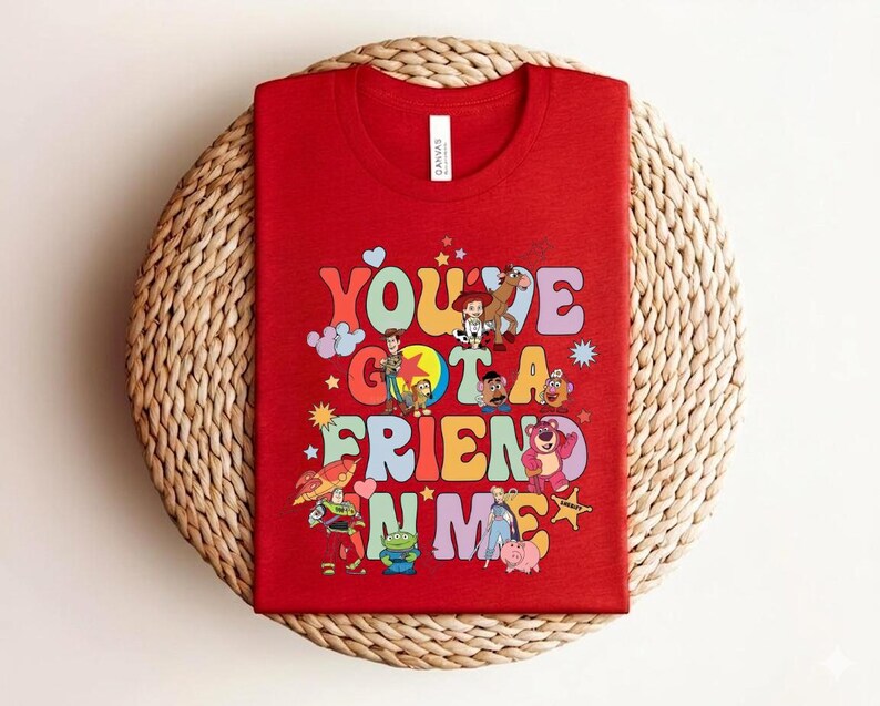 May include: Red t-shirt with the phrase "You've Got a Friend in Me" in colourful, retro lettering. The shirt features cartoon characters. The t-shirt is displayed on a woven, circular mat.