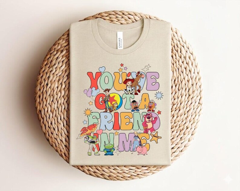May include: Beige t-shirt featuring the phrase "You've Got a Friend in Me" in a retro font, with cartoon characters. The design includes a cowboy, a space ranger, and a pink bear, reminiscent of a popular animated film.