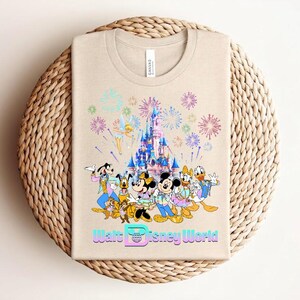 Może przedstawiać: Beżowa koszulka z kolorową grafiką postaci Disneya, zamku i fajerwerk&oacute;w. Tekst "Walt Disney World" jest wyświetlany w stylizowanej czcionce. Koszulka jest złożona na tkanej, okrągłej powierzchni.