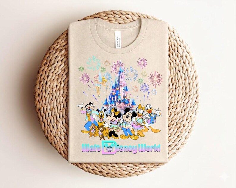 Może przedstawiać: Beżowa koszulka z kolorową grafiką postaci Disneya, zamku i fajerwerk&oacute;w. Tekst "Walt Disney World" jest wyświetlany w stylizowanej czcionce. Koszulka jest złożona na tkanej, okrągłej powierzchni.