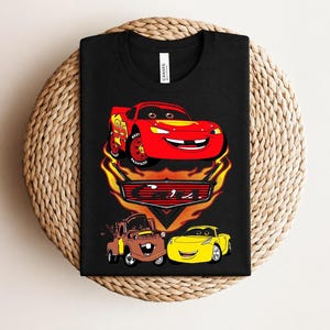 May include: Black t-shirt featuring a graphic of Lightning McQueen, Mater, and a yellow car from the movie Cars. The design includes flames and the number 95 on Lightning McQueen. The shirt is displayed on a woven surface.
