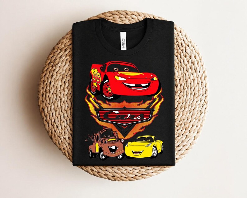 May include: Black t-shirt featuring a graphic of Lightning McQueen, Mater, and a yellow car from the movie Cars. The design includes flames and the number 95 on Lightning McQueen. The shirt is displayed on a woven surface.