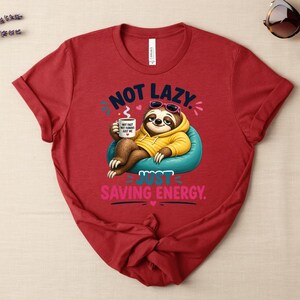 May include: Red t-shirt with the text "NOT LAZY, JUST SAVING ENERGY." The graphic features a cartoon sloth wearing sunglasses and a yellow hoodie, holding a mug. The sloth is sitting on a blue beanbag chair.