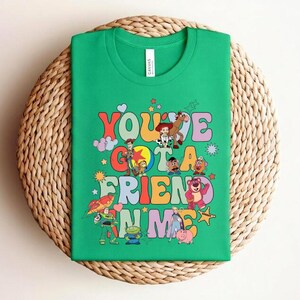 May include: A green t-shirt with the text "You've Got a Friend in Me" in a retro font, accompanied by cartoon characters. The shirt is displayed on a woven, circular surface. The t-shirt is a vibrant and playful design.