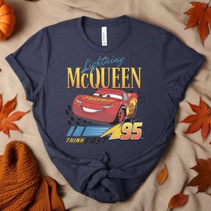 May include: Navy blue t-shirt with a graphic of Lightning McQueen from the Cars film. The design includes McQueen, the number 95, and the text "Lightning McQueen" and "Think Fast" in yellow and blue.