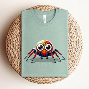 Pu&ograve; includere: T-shirt verde chiaro con un disegno di ragno dei cartoni animati. Il ragno ha una testa rossa, arancione e gialla con grandi occhi e zampe rosse e nere. La maglietta &egrave; piegata su una superficie tessuta.