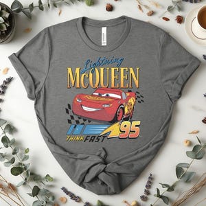 May include: A grey t-shirt with a graphic of Lightning McQueen, a red race car from the film Cars. The design includes the text "Lightning McQueen", the number "95", and the phrase "Think Fast".