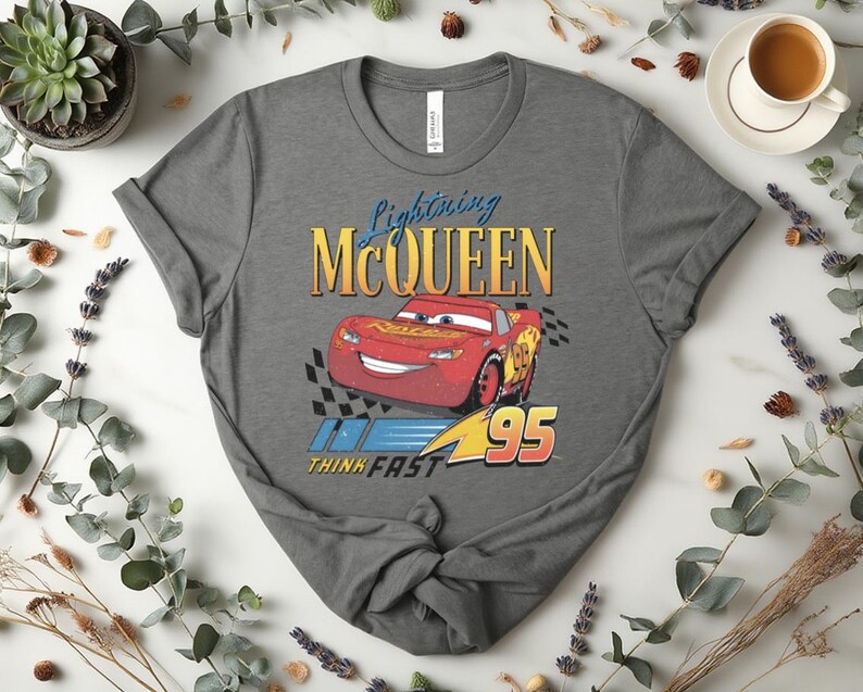 May include: A grey t-shirt with a graphic of Lightning McQueen, a red race car from the film Cars. The design includes the text "Lightning McQueen", the number "95", and the phrase "Think Fast".
