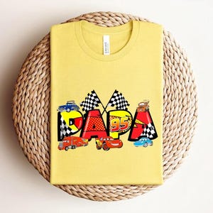 Puede incluir: Camiseta amarilla mostaza con la palabra "DADA" en un dise&ntilde;o colorido con coches de carreras y banderas a cuadros. El n&uacute;mero "95" tambi&eacute;n est&aacute; incluido. La camiseta es de un material suave.