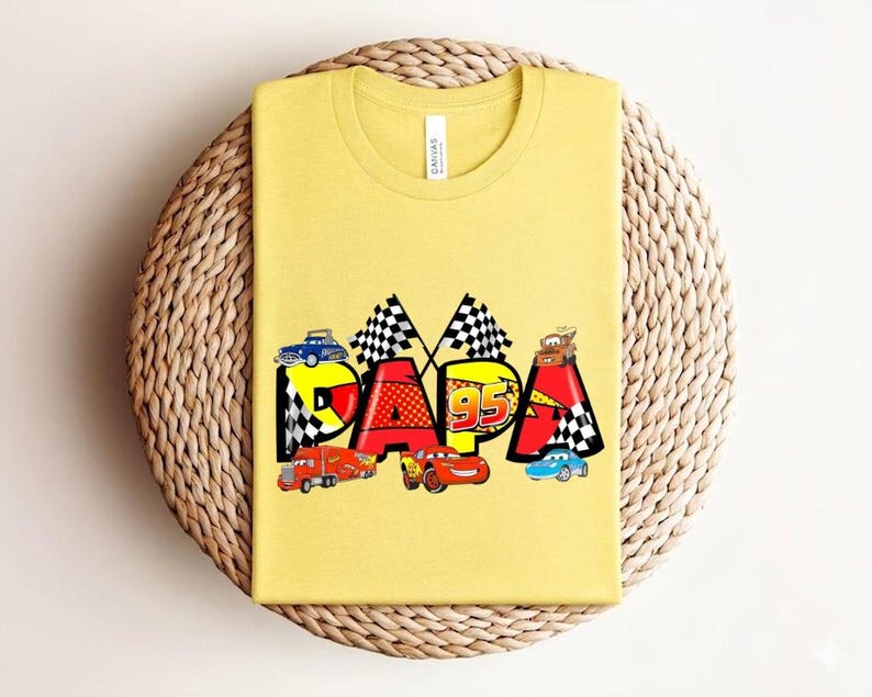 Puede incluir: Camiseta amarilla mostaza con la palabra "DADA" en un dise&ntilde;o colorido con coches de carreras y banderas a cuadros. El n&uacute;mero "95" tambi&eacute;n est&aacute; incluido. La camiseta es de un material suave.