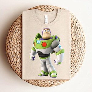 May include: A beige t-shirt featuring a colorful graphic of Buzz Lightyear from Toy Story. The character is depicted in his signature green, white, and purple spacesuit. The shirt is folded on a woven, circular surface.