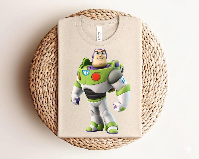 May include: A beige t-shirt featuring a colorful graphic of Buzz Lightyear from Toy Story. The character is depicted in his signature green, white, and purple spacesuit. The shirt is folded on a woven, circular surface.