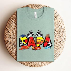 Puede incluir: Camiseta verde claro con la palabra "PAPA" en letras coloridas y coches de carreras. La "P" es azul, la "A" es roja y la segunda "P" es amarilla. La camiseta tiene cuello redondo y mangas cortas.