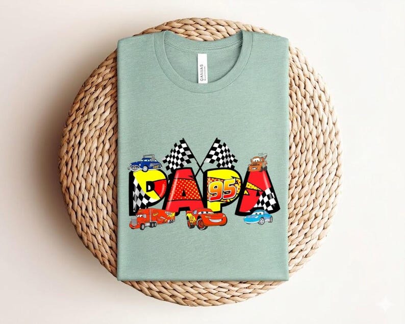 Puede incluir: Camiseta verde claro con la palabra "PAPA" en letras coloridas y coches de carreras. La "P" es azul, la "A" es roja y la segunda "P" es amarilla. La camiseta tiene cuello redondo y mangas cortas.