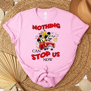 Puede incluir: Camiseta rosa claro con un dibujo animado de Mickey y Minnie Mouse en un coche rojo, con el texto "NOTHING CAN STOP US NOW" en rojo. La camiseta est&aacute; hecha de material suave.