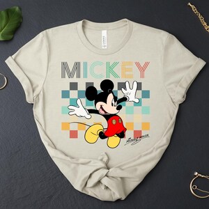 May include: Beige t-shirt featuring a colorful graphic of Mickey Mouse with the word "MICKEY" above him. Mickey is wearing his signature red shorts, yellow shoes, and white gloves. The shirt has a classic crew neck and short sleeves.