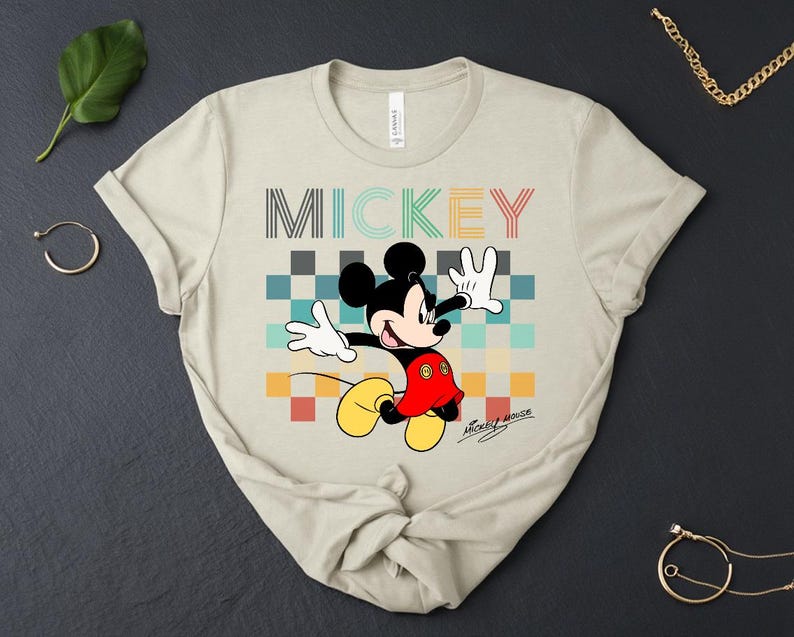 May include: Beige t-shirt featuring a colorful graphic of Mickey Mouse with the word "MICKEY" above him. Mickey is wearing his signature red shorts, yellow shoes, and white gloves. The shirt has a classic crew neck and short sleeves.