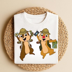 May include: White t-shirt featuring a cartoon illustration of two chipmunks dressed as explorers. They wear hats, backpacks, and hold a compass, camera, map, and flashlight. The chipmunks are brown and tan.