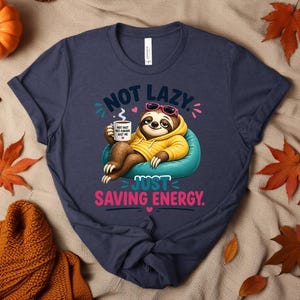 May include: Navy blue t-shirt featuring a cartoon sloth in sunglasses and a yellow hoodie, holding a coffee mug. The text on the shirt says "NOT LAZY, JUST SAVING ENERGY."