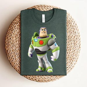 May include: Dark green t-shirt featuring a colorful graphic of Buzz Lightyear from Toy Story. The character is depicted in his signature space ranger suit with white, green, and purple accents. The shirt is displayed on a woven, circular surface.