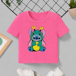 May include: A pink short-sleeved t-shirt featuring a cartoon character in shades of blue, green, and yellow. The character has large eyes and is sitting down. White sneakers are at the bottom of the image.