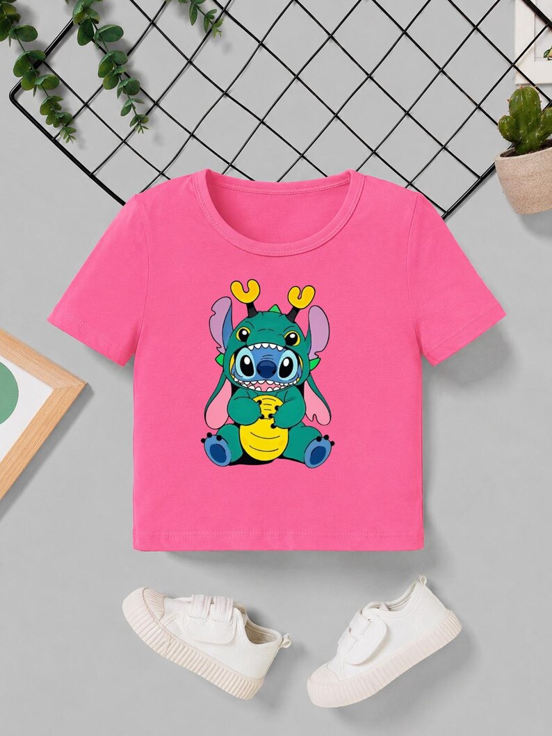 May include: A pink short-sleeved t-shirt featuring a cartoon character in shades of blue, green, and yellow. The character has large eyes and is sitting down. White sneakers are at the bottom of the image.
