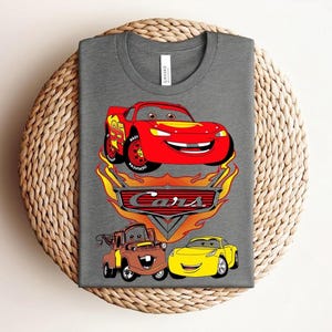 May include: A gray t-shirt featuring a colorful graphic of characters from the animated movie "Cars." The design includes a red race car, a brown tow truck, and a yellow car, with the "Cars" logo and flames.