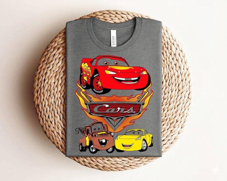 May include: A gray t-shirt featuring a colorful graphic of characters from the animated movie "Cars." The design includes a red race car, a brown tow truck, and a yellow car, with the "Cars" logo and flames.