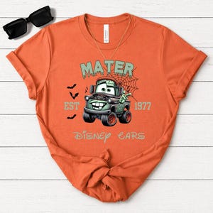 Może przedstawiać: Pomarańczowa koszulka z nadrukiem postaci Mater z filmu Auta Disneya, stylizowanego na potwora Frankensteina. Projekt zawiera napisy "MATER", "EST 1977" i "DISNEY CARS", z nietoperzami i pajęczyną. Koszulka ma okrągły dekolt.