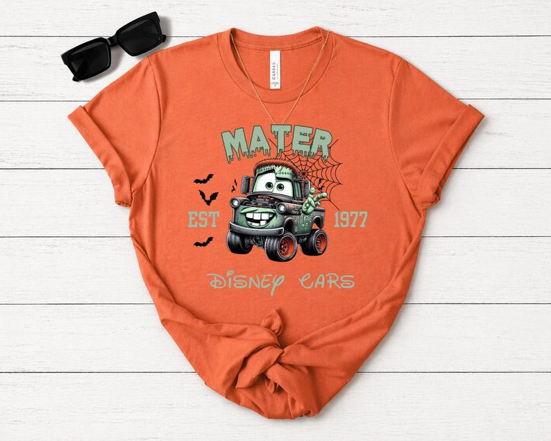 Może przedstawiać: Pomarańczowa koszulka z nadrukiem postaci Mater z filmu Auta Disneya, stylizowanego na potwora Frankensteina. Projekt zawiera napisy "MATER", "EST 1977" i "DISNEY CARS", z nietoperzami i pajęczyną. Koszulka ma okrągły dekolt.
