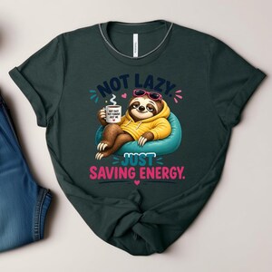 May include: Dark green t-shirt with a cartoon sloth wearing sunglasses and a yellow hoodie, relaxing on a blue beanbag chair. The shirt has the text "NOT LAZY JUST SAVING ENERGY" and a coffee cup graphic.